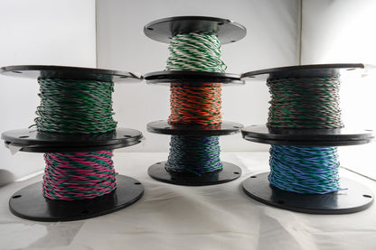 Three large black spools of braided wire or cable, each wound with a different color combination: green and white, orange and black, and pink and blue, displayed on a white surface against a white background.