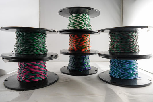 Three large black spools of braided wire or cable, each wound with a different color combination: green and white, orange and black, and pink and blue, displayed on a white surface against a white background.